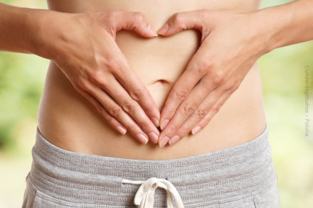 Eine Person formt mit ihren Händen ein Herz über ihrem nackten Bauch. Die Person trägt eine graue Hose mit weißem Kordelzug. Der Hintergrund ist unscharf und in natürlichen Farben gehalten.