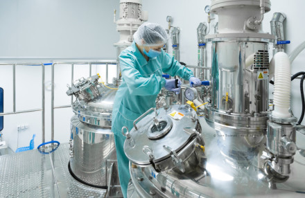Eine Person in Schutzkleidung, einschließlich Haarnetz, Maske und Handschuhen, arbeitet in einer modernen Produktionsanlage mit glänzenden Edelstahlbehältern und Maschinen.