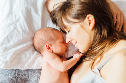 Eine Frau liegt auf einem Bett und hält ein neugeborenes Baby eng an ihrem Gesicht. Beide haben die Augen geschlossen und sehen friedlich aus. Die Frau hat lange braune Haare, und das Baby ist nackt. Sie liegen auf weißen Laken und einer grauen Decke.