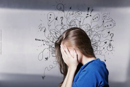 Eine Frau hält sich den Kopf in den Händen und wirkt gestresst. Hinter ihr sind viele chaotische Kritzeleien, wie Glühbirnen, Ausrufezeichen und Fragezeichen, gezeichnet, die ihren unruhigen Geisteszustand symbolisieren. Sie trägt ein blaues Oberteil.