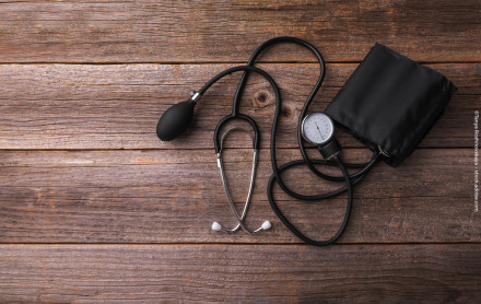 Auf dem Bild ist ein Stethoskop und ein Blutdruckmessgerät auf einer hölzernen Oberfläche zu sehen. Das Blutdruckmessgerät hat eine schwarze Manschette und eine schwarze Birne, die mit dem Messgerät verbunden sind.