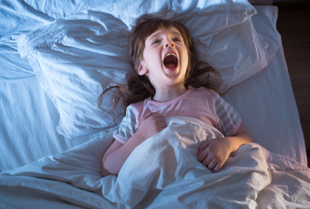 Ein kleines Mädchen liegt in einem Bett und scheint erschrocken oder verängstigt zu sein. Sie hat den Mund weit geöffnet und schreit. Sie trägt ein rosa Schlafanzug-Oberteil und hält die Bettdecke fest. Die Bettwäsche und das Kopfkissen sind weiß.