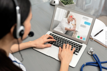 Eine Person mit einem Headset führt eine Telemedizin-Beratung über ihren Laptop durch. Auf dem Laptop-Bildschirm ist eine ältere Frau zu sehen, die anscheinend Kopfschmerzen hat und ihr Kopf mit ihrer Hand stützt.
