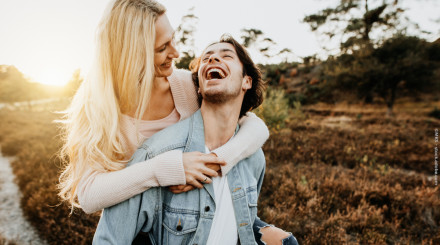 Ein lächelnder Mann trägt eine blonde Frau auf dem Rücken, die ihn liebevoll umarmt. Beide sind glücklich und genießen die Natur im Hintergrund, die von Bäumen und einem Weg geprägt ist. Das Sonnenlicht scheint warm herein.