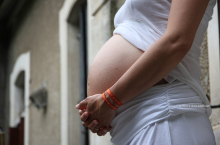 Eine schwangere Frau, die ein weißes Shirt und eine weiße Hose trägt und von der Seite fotografiert wird. Sie hält ihre Hände vor ihrem Bauch zusammengefaltet. Im Hintergund ist ein unscharfes Gebäude zu sehen.
