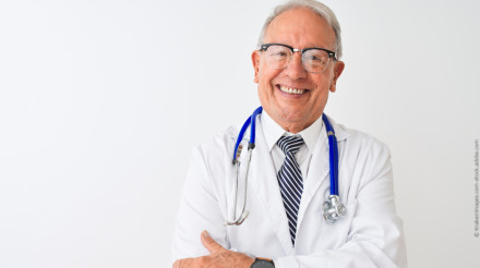 Ein älterer Arzt mit grauem Haar und Brille trägt einen weißen Kittel und ein Stethoskop um den Hals und lächelt in die Kamera. Er steht vor einem weißen Hintergrund und hat die Arme verschränkt.