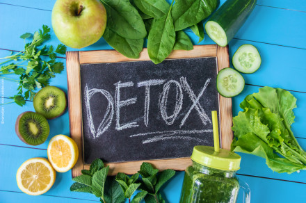 Tafel mit der Aufschrift "DETOX" umgeben von frischem Obst und Gemüse, darunter ein grüner Apfel, Spinatblätter, Gurkenscheiben, Blattsalat, Kiwischeiben, Zitronenscheiben und ein Smoothie in einem Glas mit gelbem Deckel und Strohhalm.