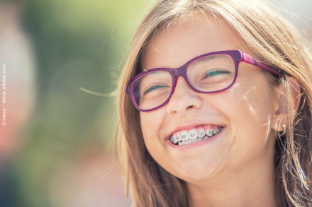 Ein lächelndes Mädchen mit einer Zahnspange und lila Brille. Hintergrund unscharf mit grünlichen Tönen. Gesichtsnahaufnahme bei Tageslicht.