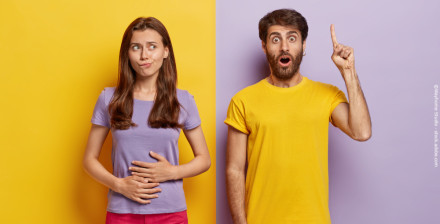 Ein junges Paar steht vor einer zweifarbigen Wand, die in Gelb und Lila geteilt ist. Die Frau links trägt ein lila T-Shirt und fasst sich mit besorgtem, überlegendem Gesichtsausdruck an den Bauch.