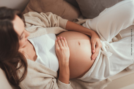 Eine schwangere Frau liegt auf dem Sofa und hält sanft ihren nackten Babybauch mit ihren Händen. Sie trägt bequeme, helle Kleidung.