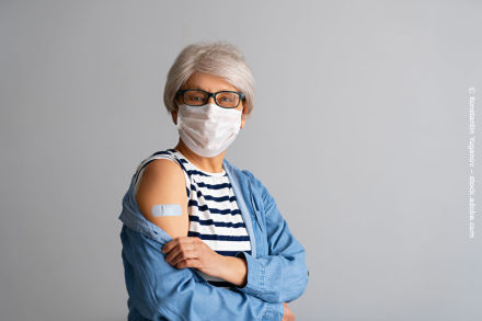 Ältere Person mit weißem Haar und Brille trägt eine Gesichtsmaske und zeigt ihren Arm mit einem Pflaster nach einer Impfung. Sie trägt ein gestreiftes Oberteil und ein blaues Hemd. Hintergrund ist grau.
