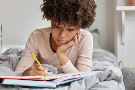 Eine junge Frau liegt auf dem Bett und schreibt in ein Notizbuch. Sie trägt ein weißes Oberteil und hält einen gelben Stift in der Hand. Ihr Gesichtsausdruck ist konzentriert, und sie stützt ihren Kopf mit einer Hand ab. Der Hintergrund ist unscharf.
