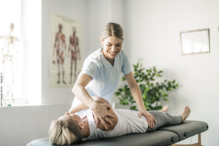 Eine Physiotherapeutin behandelt eine liegende Patientin auf einer Therapieliege. Die Therapeutin lächelt, während sie an der Schulter ihrer Patientin arbeitet. Im Hintergrund sind ein anatomisches Poster und eine Pflanze zu sehen.