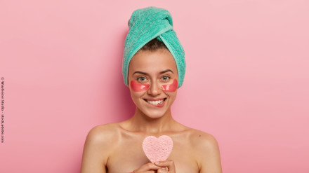 Eine lächelnde Frau mit einer türkisfarbenen Handtuch-Turban auf dem Kopf hat rosa Augenpads unter ihren Augen und hält einen herzförmigen Schwamm. Sie steht vor einem rosa Hintergrund.