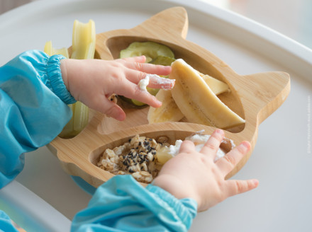 Nahaufnahme von Babyhänden, die auf einem Baby-Esstisch eine bambusförmige Teller mit Bananenscheiben, Gurken, Selleriestangen und Haferflocken mit Sesamkernen berühren. Das Baby trägt einen blauen Ärmel.