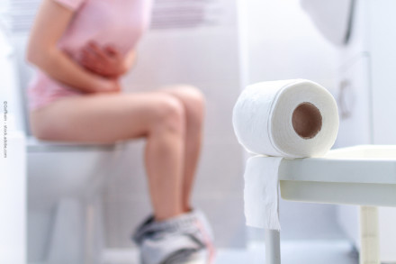 Eine Person sitzt auf einer Toilette und hält sich den Bauch. Es gibt eine nahe Ansicht einer Toilettenpapierrolle im Vordergrund. Die Hose der Person ist heruntergelassen und liegt um ihre Knöchel.