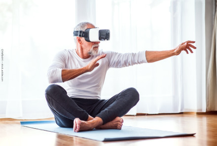 Ein älterer Mann mit grauem Haar und Bart sitzt im Schneidersitz auf einer Yogamatte in einem hellen Raum. Er trägt eine Virtual-Reality-Brille und hat die Arme ausgestreckt, als ob er eine Interaktion in der virtuellen Welt erlebt.