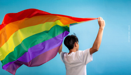 Eine Person mit kurzen, dunklen Haaren steht vor einem blauen Hintergrund und hält eine große Regenbogenflagge, die im Wind flattert, über ihrem Kopf. Die Person trägt ein weißes T-Shirt und ist mit dem Rücken zur Kamera gewandt.