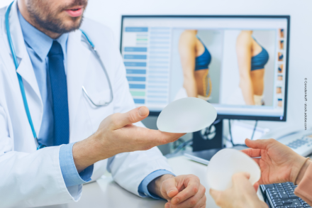 Ein Arzt im weißen Kittel zeigt einer Patientin zwei Silikonimplantate. Im Hintergrund ist ein Monitor mit Vorher-Nachher-Bildern einer Brustvergrößerung zu sehen.