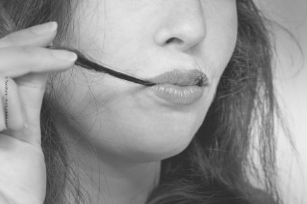 Schwarz-Weiß-Nahaufnahme einer Person, die eine Strähne ihres Haares mit den Fingern hält und es mit den Lippen berührt. Die Lippen sind leicht geschlossen und glänzend. Ein Teil des Gesichts und der Haare ist sichtbar.