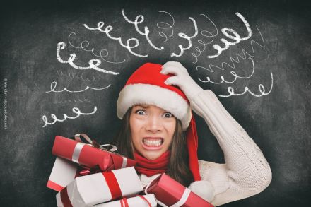 Eine Frau trägt eine Weihnachtsmütze und hält mehrere Geschenke in den Händen. Sie sieht gestresst aus und hat ein verängstigtes Gesicht. Im Hintergrund ist eine Tafel mit Kritzeleien zu sehen, die Verwirrung und Stress symbolisieren.