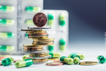 Ein Stapel Euro-Münzen mit einer Ein-Cent-Münze an der Spitze, umgeben von grünen und weißen Kapseln. Im Hintergrund sind unscharfe Blisterpackungen mit ähnlichen Kapseln zu sehen.