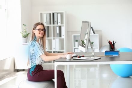 Eine lächelnde Frau sitzt auf einem Gymnastikball an einem Schreibtisch. Sie trägt eine Brille und tippt auf einem Computer. Neben ihr stehen eine Tasse, Notizbücher und Stifte. Im Hintergrund sind Regale mit Ordnern und eine Pflanze.