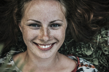 Nahaufnahme eines lächelnden Gesichts einer Frau mit hellen Augen, Sommersprossen und langen braunen Haaren. Sie liegt auf dem Rücken im Gras und trägt ein buntes Oberteil.