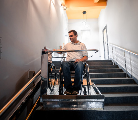 Ein Mann im Rollstuhl benutzt einen Treppenlift, um eine Treppe im Inneren eines Gebäudes hinunterzufahren. Er trägt ein weißes Polohemd und blaue Jeans. Die Treppe hat ein Metallgeländer auf beiden Seiten und die Wände sind hell gestrichen.