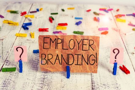 Ein Schild aus braunem Papier mit dem handgeschriebenen Text "EMPLOYER BRANDING!" steht auf einer weiß verwitterten Holzoberfläche, die mit bunten Wäscheklammern dekoriert ist.