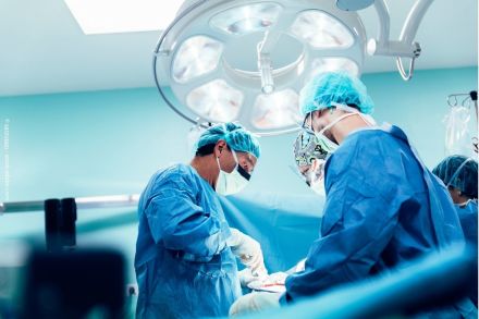 Mehrere Chirurgen in blauen OP-Kitteln und Mundschutz bei einer Operation im Operationssaal, beleuchtet durch eine große OP-Lampe.