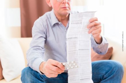 Ein älterer Mann sitzt auf einem Sofa und liest die Packungsbeilage eines Medikaments. In der rechten Hand hält er eine Blisterpackung mit Tabletten. Er trägt ein gestreiftes Hemd und eine Brille. Im Hintergrund sind eine Couch und ein Vorhang zu sehen.