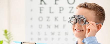 Ein Junge steht vor einer Tafel mit unterschiedlich großen Buchstaben und hat eine Brille vom Augenarzt auf.