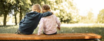 Zwei Kinder sitzen mit auf einer Bank und blicken auf eine Wiese. Der kleine Junge legt den Arm um seine Schwester.