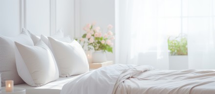Helles, gemütliches Schlafzimmer mit einem aufgeräumten Bett, weichen weißen Kissen und einer weißen Bettdecke. Im Hintergrund fällt sanftes Tageslicht durch ein großes Fenster herein, das von weißen Vorhängen umrahmt wird.