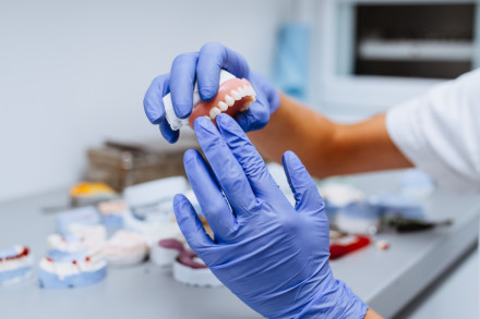 Arbeit an einer Zahnprothese. Hände in blauen Handschuhen überprüfen eine fertige Zahnprothese für den Oberkiefer. Im Hintergrund weitere Arbeitsstücke.