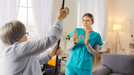 Eine wütende ältere Patientin erhebt ihren Stock und bedroht damit eine Krankenschwester in einem Pflegeheim. Eine angespannte Konfliktsituation, die emotionale Not, medizinische Herausforderungen und Schwierigkeiten bei der Pflege widerspiegelt.