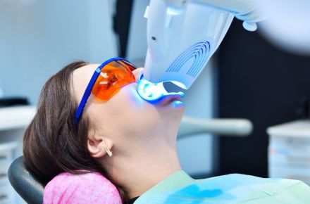 Eine Frau bleacht ihre Zähne, sie hält dafür ein kleines Bleachinggerät mit blauem Licht an ihren Mund.