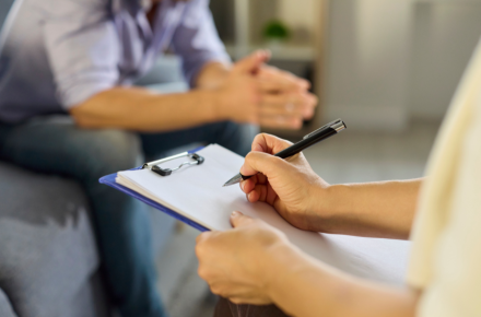 Therapeutin macht Notizen auf einem Klemmbrett während eines Gesprächs mit einem Patienten in einer Beratungssituation zur Burnout-Behandlung und Stressbewältigung in einer ruhigen Praxisumgebung.