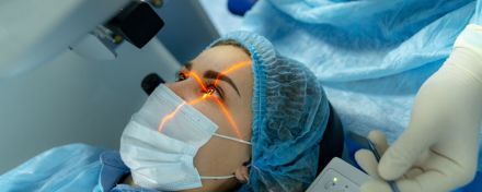 Eine Frau liegt auf einem Behandlungsstuhl und trägt OP-Kleidung. Ihr wird mit einem orangenen Laser in das Auge geleuchtet. 