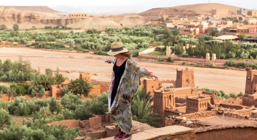 The girl-traveler in a hat is standing on a high wall with arms outstretched in the background of the city in the desert of Ait BenHaddou, Morocco, Africa