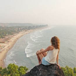 A young girl with blond hair sits on top of a mountain and looks at the sea and beach, Arambol, Goa, India