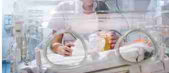 A healthcare professional tending to a baby in an incubator in a neonatal unit. The adult is wearing glasses and a white uniform, and the scene suggests a hospital environment with medical equipment visible.