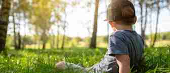 A young child wearing a black cap and grey shirt sits on the green grass in a sunlit forest. The child faces away, looking towards trees with sunlight filtering through the leaves, creating a serene and peaceful atmosphere.