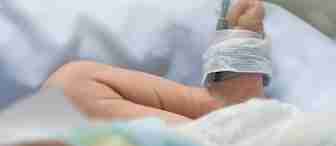 Close-up of a newborn baby's foot with a medical sensor wrapped around it, lying on a white sheet. The baby's leg is slightly bent, and the background is softly blurred, creating a gentle, calm atmosphere typical of a hospital nursery or neonatal unit.