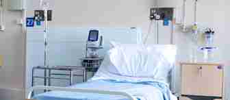 A hospital room with a single bed, white pillows, and light blue sheets. An IV stand and medical equipment are positioned next to the bed. A water jug and glass rest on a small wooden bedside table. The room appears clean and orderly.