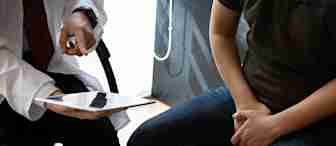 A doctor in a white coat is showing information on a digital tablet to a patient. The patient is sitting with their hands on their lap, slightly hunched forward, and appears to be attentively listening. The setting looks like a medical consultation room.