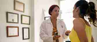 A doctor in a white coat is speaking with a patient dressed in a yellow outfit in a medical office. The doctor is holding a sphygmomanometer. Certificates are hung on the wall behind them, and medical equipment is visible in the background.
