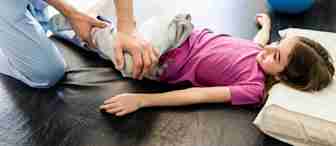 A physical therapist helps a young girl lying on a table by gently stretching her leg. The girl is wearing a pink shirt and gray pants, and she rests her head on a pillow. Exercise equipment is seen in the background.