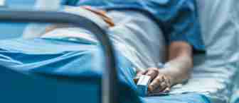 A person lies in a hospital bed wearing a blue gown, with one hand resting beside them holding a medical finger clip, possibly a pulse oximeter. The focus is on the lower part of their body, highlighting the medical equipment and hospital surroundings.
